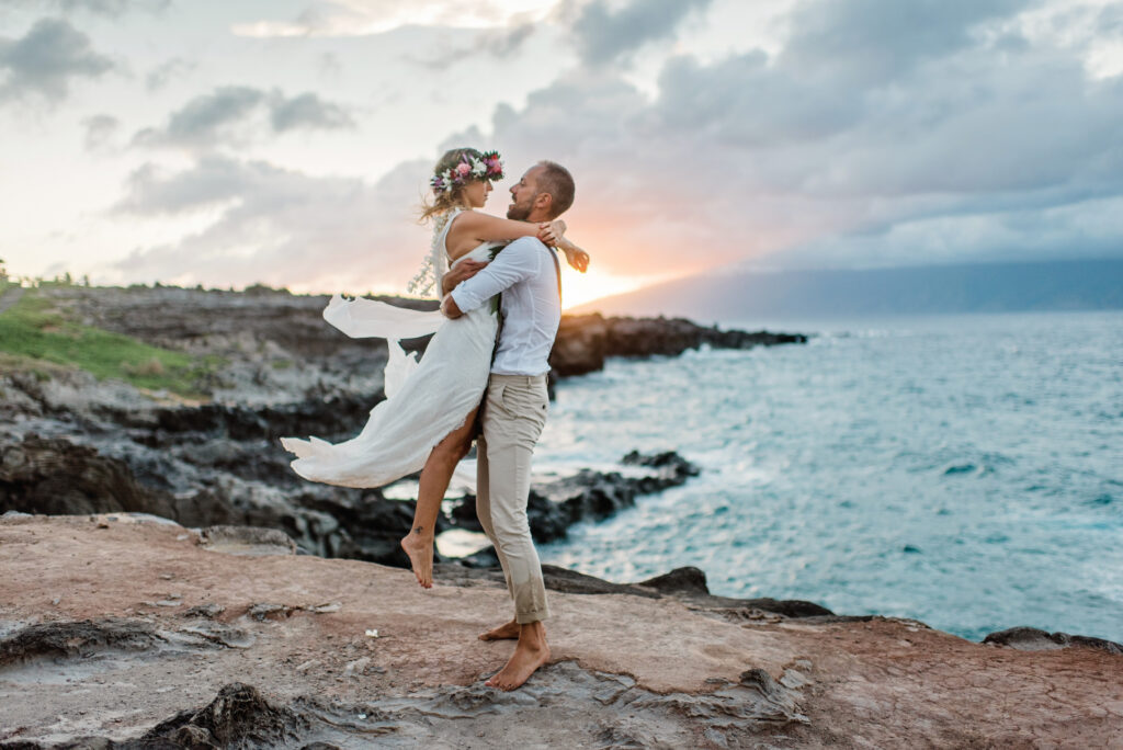 Groom lifting his bride during a sunset celebration on Maui's dramatic oceanfront coastline.