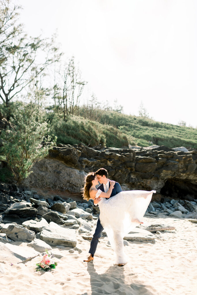 Groom carrying his bride across a Maui beach after their intimate wedding ceremony.