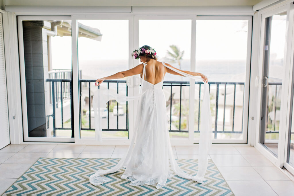 Bride in her wedding dress standing in front of ocean-view windows before a Maui elopement.