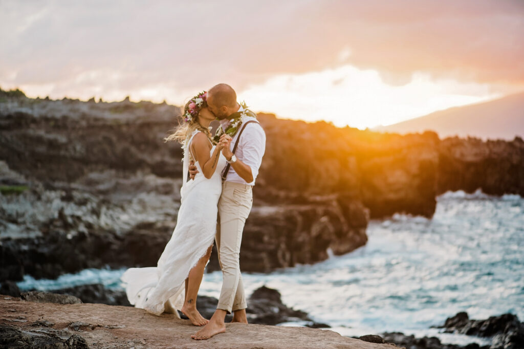 Bride and groom sharing a kiss on Maui's lava rock coastline during sunset after their beach wedding ceremony.