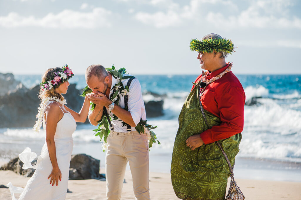 Groom kissing the bride's hand during a Maui beach wedding ring exchange with their officiant nearby.