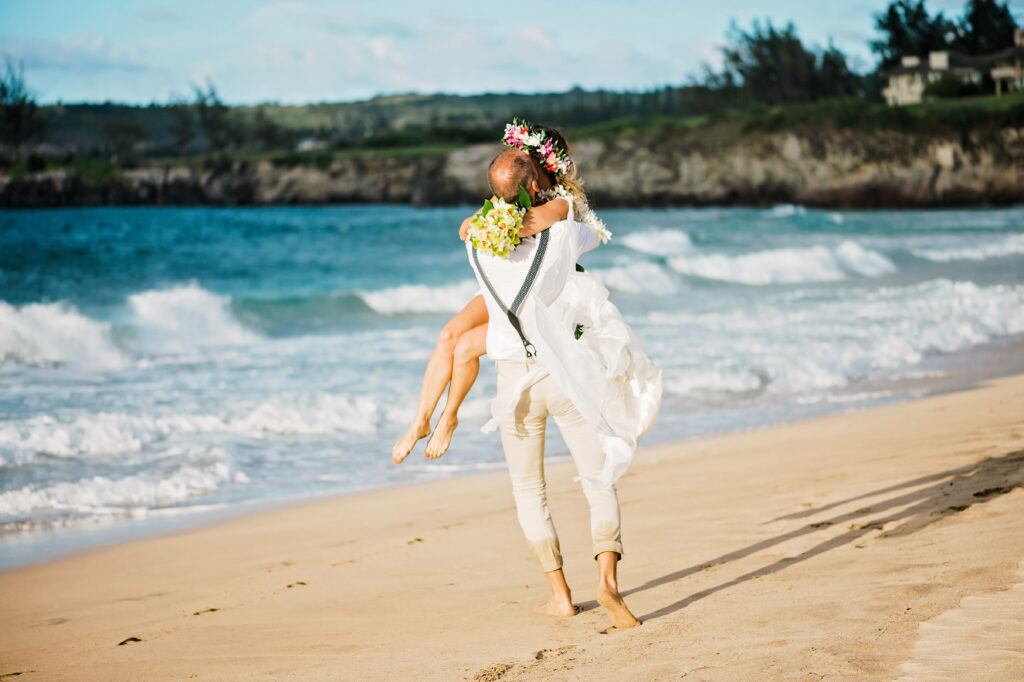 Newlywed couple embracing and walking along the shoreline after their Maui beach wedding ceremony.